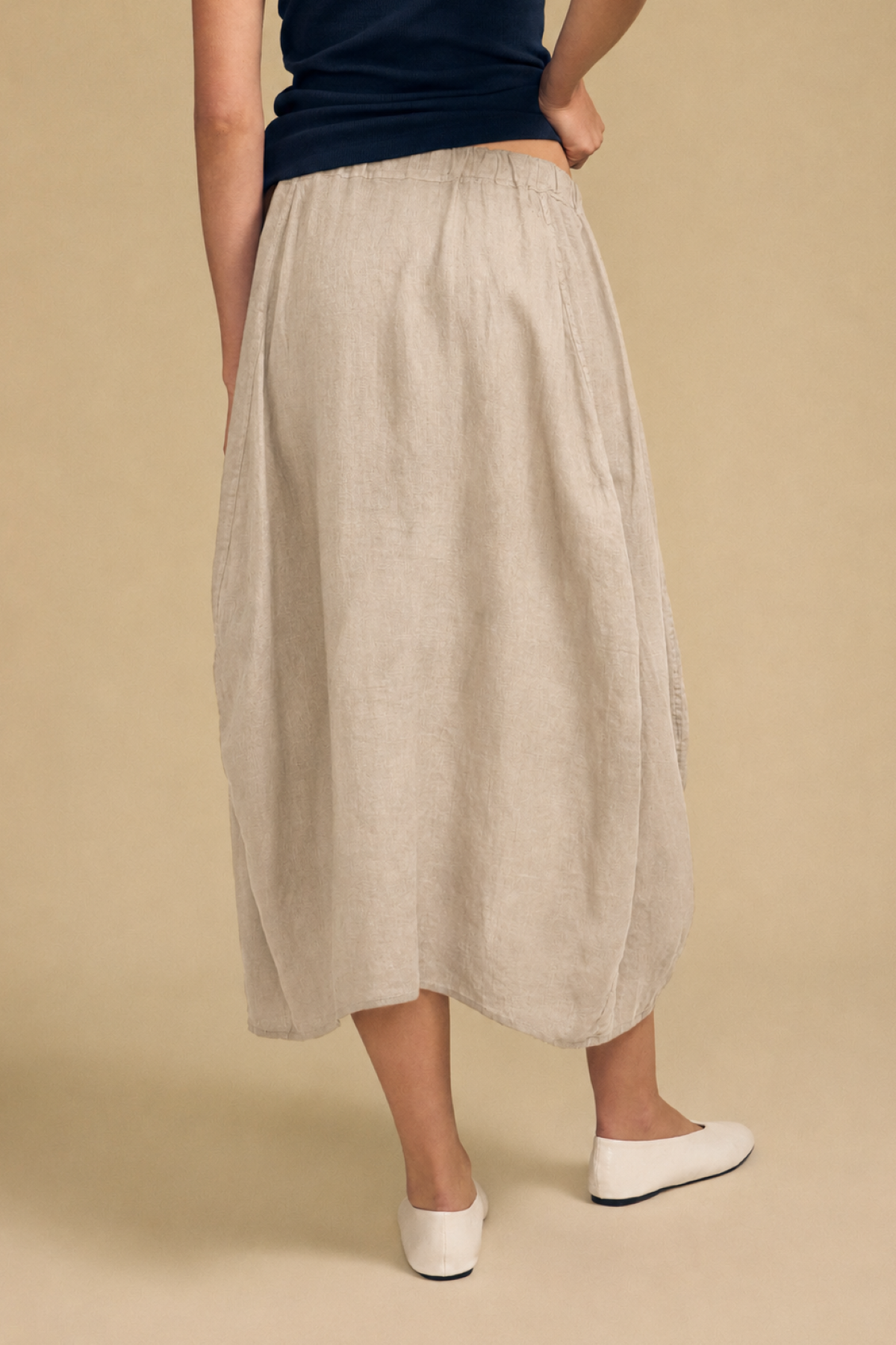 A person is shown from the back, wearing a dark top and the FAE LINEN SKIRT by Velvet by Graham & Spencer—a light beige, A-line midi skirt with an elastic waistband—paired with white flat shoes, standing against a plain beige background.-42003448922305