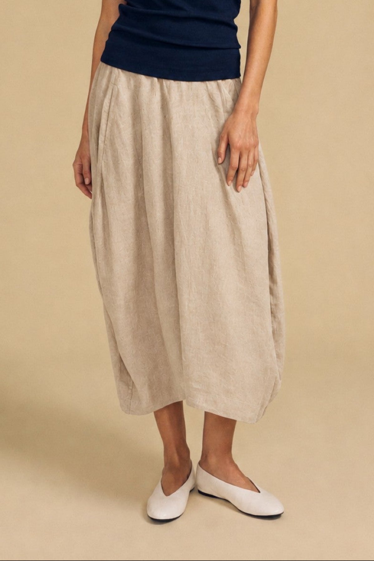 A woman stands against a beige backdrop wearing a navy sleeveless top, the Velvet by Graham & Spencer FAE LINEN SKIRT—a beige linen maxi skirt featuring an elastic waistband and A-line silhouette—and white slip-on shoes.