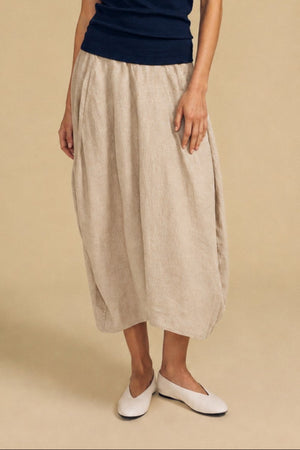 A woman stands against a beige backdrop wearing a navy sleeveless top, the Velvet by Graham & Spencer FAE LINEN SKIRT—a beige linen maxi skirt featuring an elastic waistband and A-line silhouette—and white slip-on shoes.