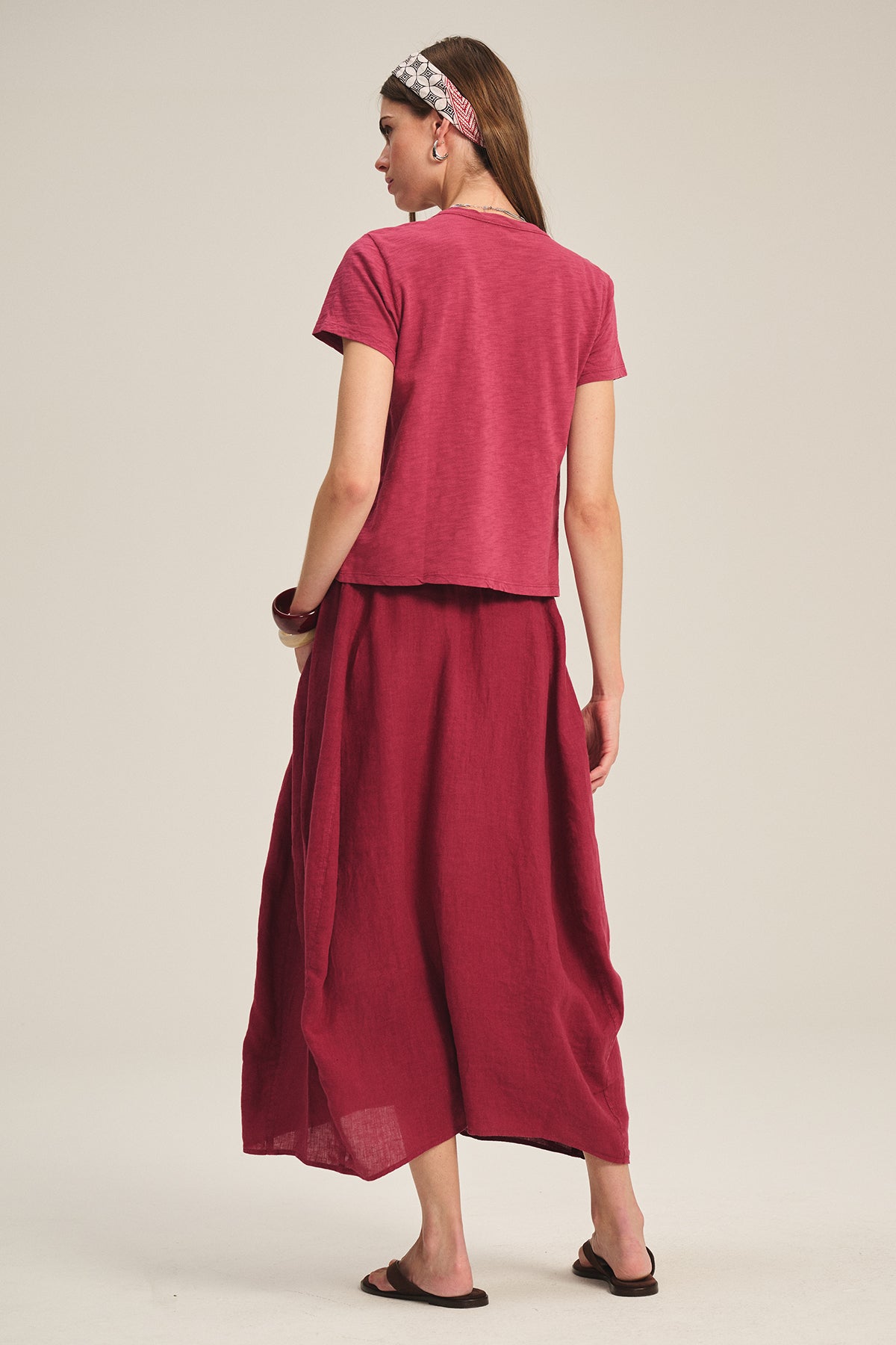 A woman stands with her back to the camera, wearing a short-sleeved red top, black sandals, a patterned headband, and the FAE LINEN SKIRT by Velvet by Graham & Spencer—a long A-line skirt with an elastic waistband.-Hover Image