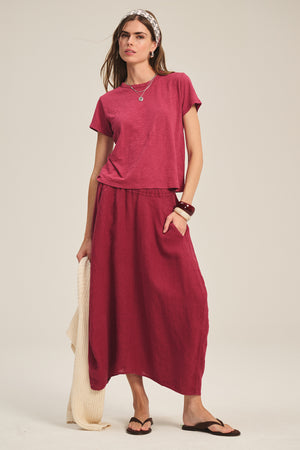 A woman wears the Velvet by Graham & Spencer FAE LINEN SKIRT, a short-sleeve maroon tee, sandals, a headband, jewelry, and holds a cream textured bag.