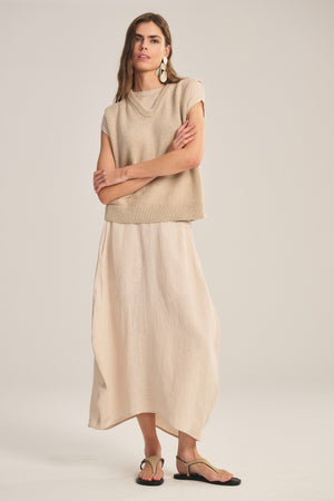 A woman stands with arms crossed in a beige short-sleeve knit top, black sandals, large earrings, and the FAE LINEN SKIRT by Velvet by Graham & Spencer, featuring spring-weight linen and an elastic waistband, against a plain light background.