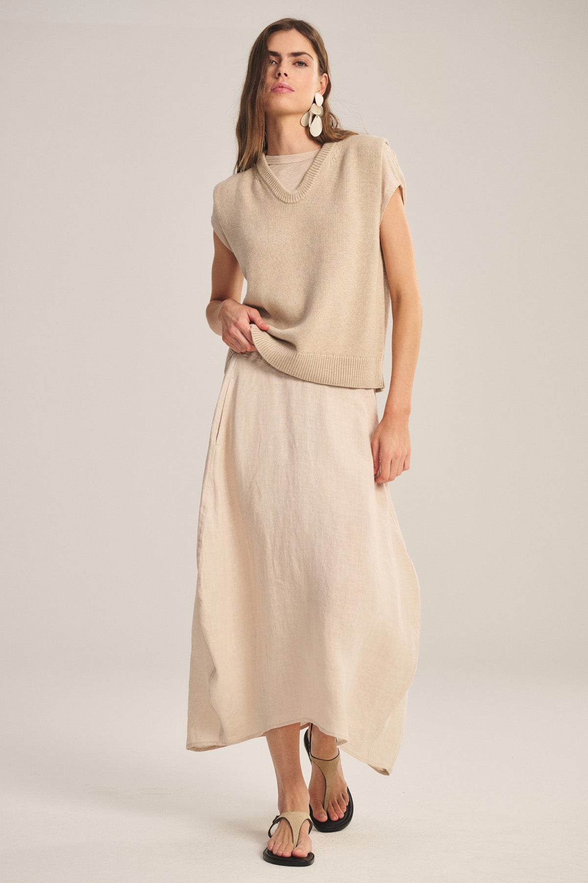 A woman wears the FAE LINEN SKIRT by Velvet by Graham & Spencer—a spring-weight, A-line skirt with an elastic waistband—paired with a sleeveless beige sweater, large white earrings, and sandals against a plain background.-41008620503233