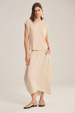 A woman wears the FAE LINEN SKIRT by Velvet by Graham & Spencer—a beige, A-line, spring-weight linen skirt with an elastic waist and side pockets—paired with a matching loose top, flat sandals, and large earrings.