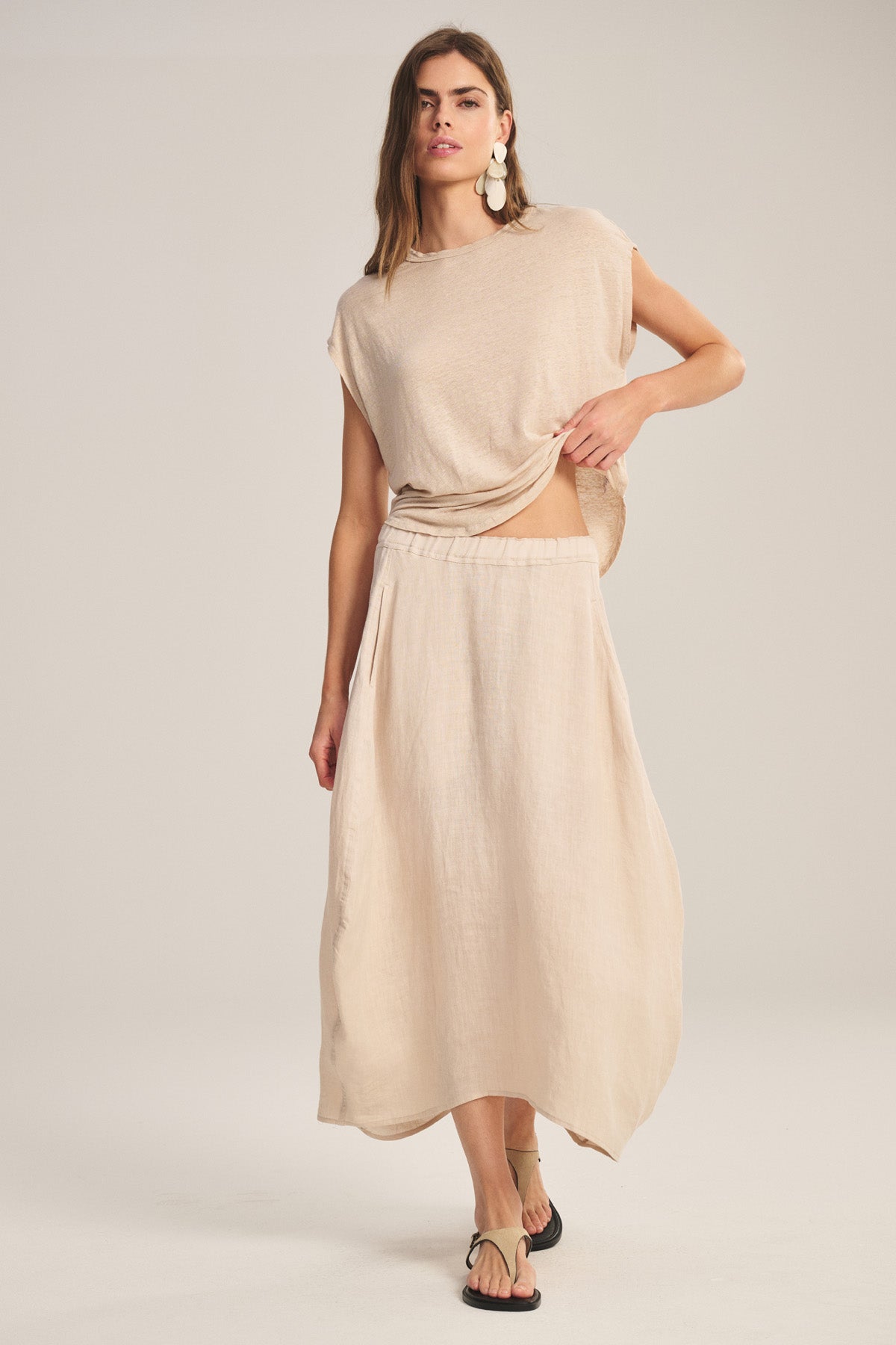 A woman stands against a plain background, slightly lifting her top while wearing the FAE LINEN SKIRT by Velvet by Graham & Spencer, styled with a loose beige tee, black sandals, and large earrings.-41008267264193