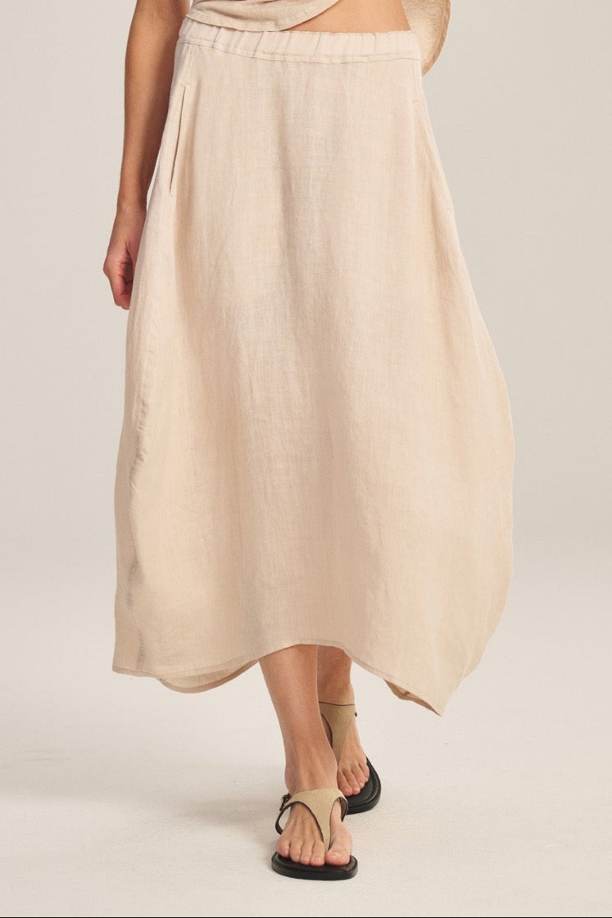 A woman stands against a plain background, slightly lifting her top while wearing the FAE LINEN SKIRT by Velvet by Graham & Spencer, styled with a loose beige tee, black sandals, and large earrings.-41008267264193
