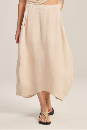 A woman stands against a plain background, slightly lifting her top while wearing the FAE LINEN SKIRT by Velvet by Graham & Spencer, styled with a loose beige tee, black sandals, and large earrings.