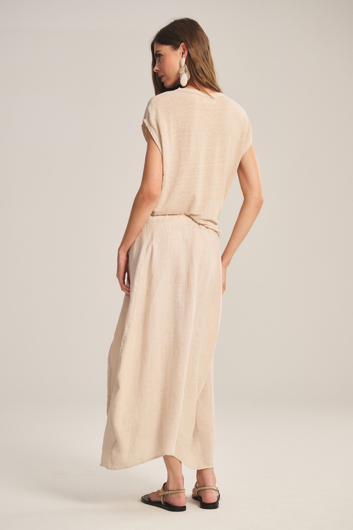 Woman stands with her back to the camera, wearing the FAE LINEN SKIRT by Velvet by Graham & Spencer, a loose beige top, black sandals, and large earrings against a plain light background.