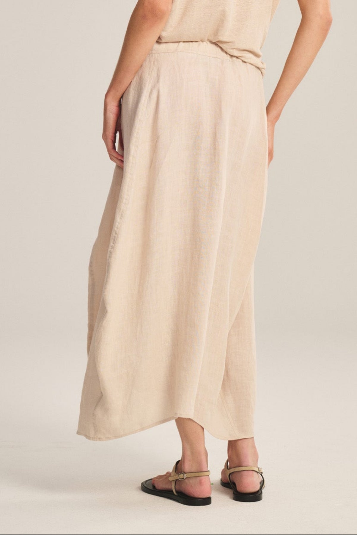 Woman stands with her back to the camera, wearing the FAE LINEN SKIRT by Velvet by Graham & Spencer, a loose beige top, black sandals, and large earrings against a plain light background.