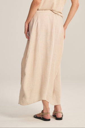 Woman stands with her back to the camera, wearing the FAE LINEN SKIRT by Velvet by Graham & Spencer, a loose beige top, black sandals, and large earrings against a plain light background.