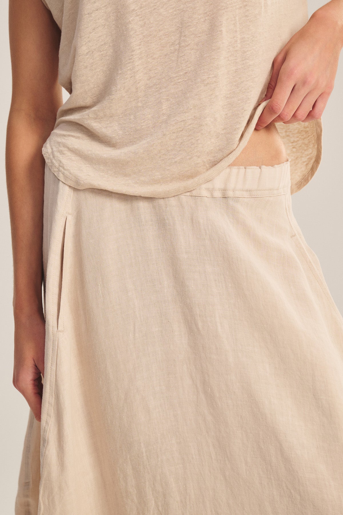 Model wears the FAE LINEN SKIRT by Velvet by Graham & Spencer, a beige A-line skirt with elastic waistband, paired with a loose matching top. Mid-torso to knee view, one hand gently holding the waistband.-41008267198657