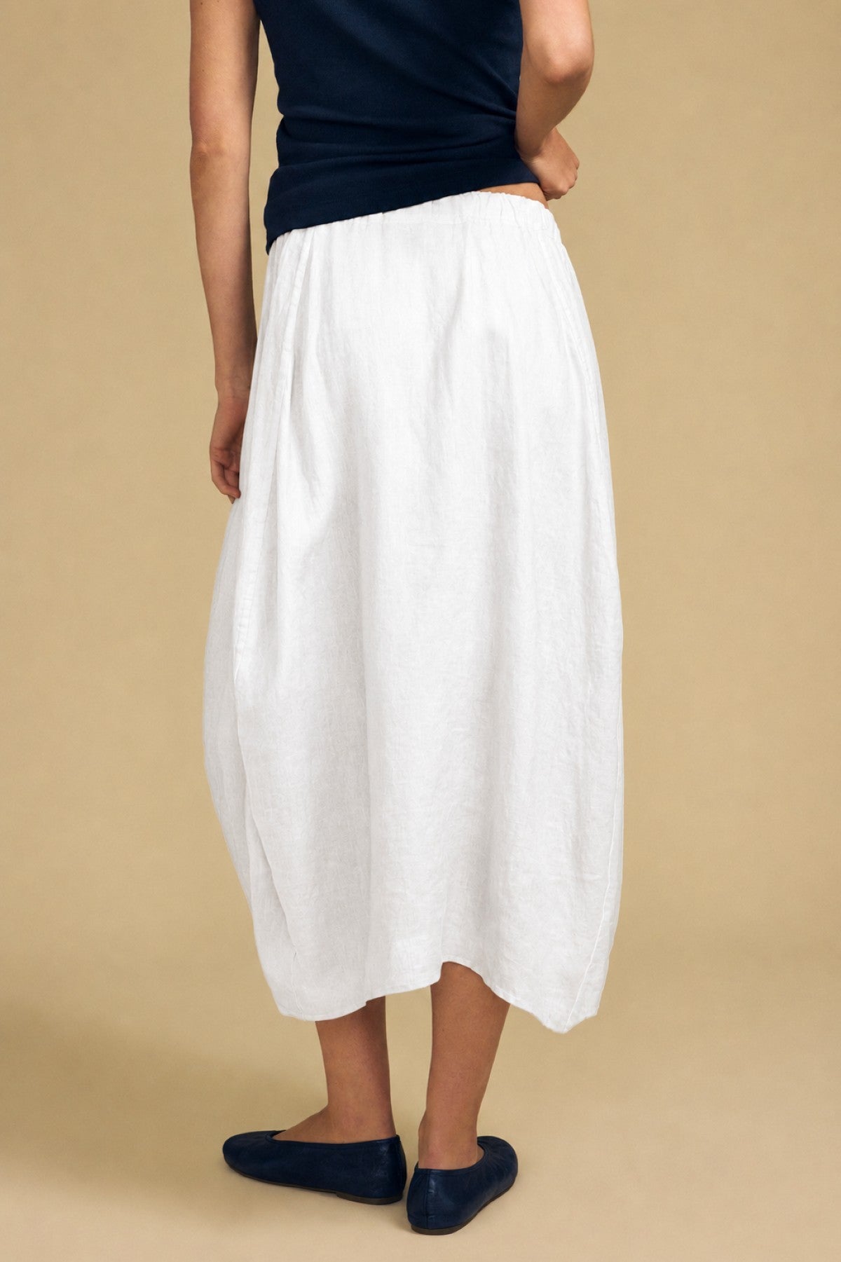 Person wearing a black top, the FAE LINEN SKIRT by Velvet by Graham & Spencer—a white linen maxi skirt featuring an A-line silhouette and elastic waistband—and black flats, standing against a beige background with their back slightly turned.