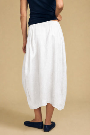 Person wearing a black top, the FAE LINEN SKIRT by Velvet by Graham & Spencer—a white linen maxi skirt featuring an A-line silhouette and elastic waistband—and black flats, standing against a beige background with their back slightly turned.