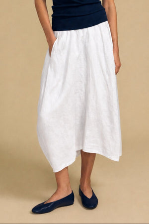 Against a tan background, a person wears the Velvet by Graham & Spencer FAE LINEN SKIRT—a white linen maxi with an elastic waistband and A-line silhouette—paired with a navy sleeveless top and navy flats, hands in pockets and gazing slightly upward.