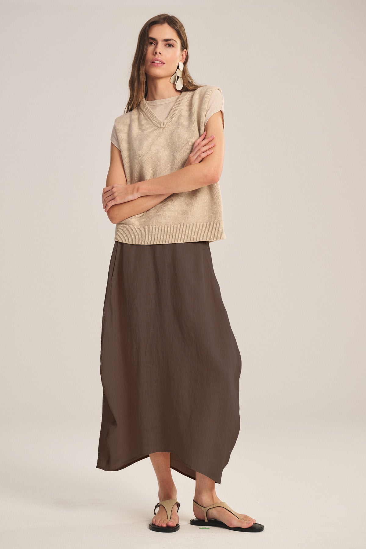 A woman stands with arms crossed, wearing a beige sleeveless top, flat sandals, large earrings, and the FAE LINEN SKIRT by Velvet by Graham & Spencer—a brown A-line linen skirt with an elastic waistband—against a plain light background.-41038318665921