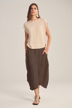 A woman wearing a beige short-sleeve top, the Velvet by Graham & Spencer FAE LINEN SKIRT—a brown A-line midi with an elastic waistband—sandals, and large white earrings stands against a plain light background.