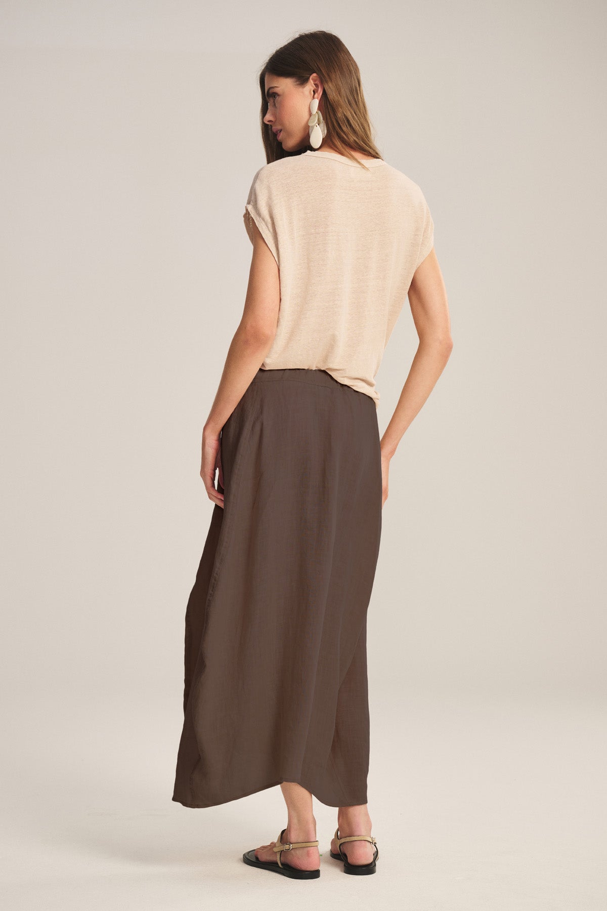 A woman faces away, wearing the FAE LINEN SKIRT by Velvet by Graham & Spencer—a long brown A-line skirt with an elastic waistband—paired with a beige short-sleeve top, flat sandals, and large drop earrings against a plain background.