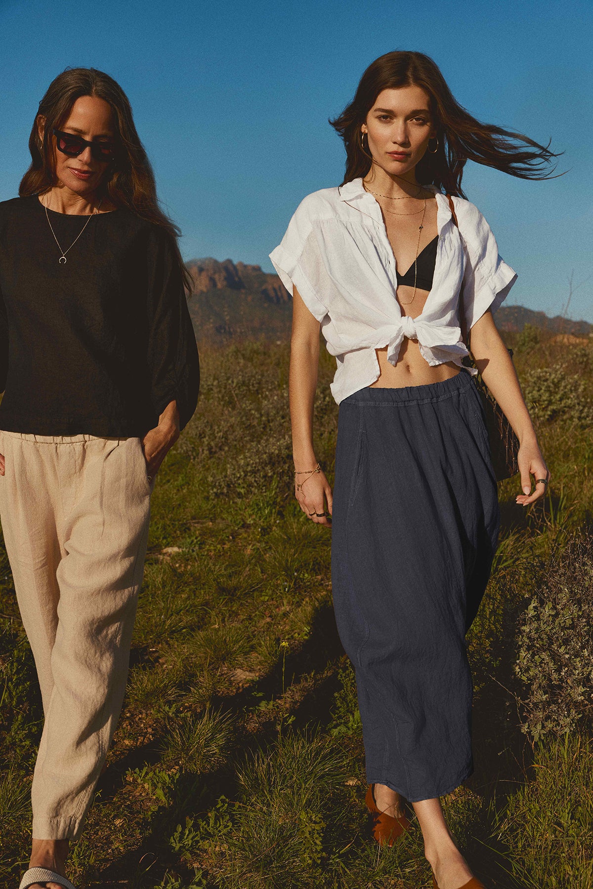   Two women walking outdoors, one dressed in a black shirt and beige pants, the other in a white cropped top and a Velvet by Graham & Spencer FAE LINEN A-LINE SKIRT with an elastic waistband, with a natural backdrop. 
