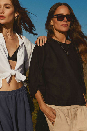 Two women are outside; one wears a white twist-knot shirt with blue pants, the other sports the CASS LINEN TOP by Velvet by Graham & Spencer in black with beige pants. Both have long hair and sunglasses.