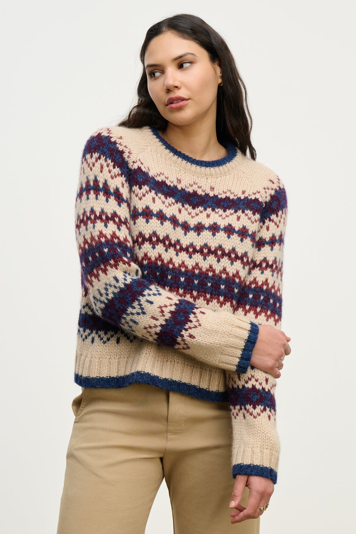 A woman stands against a plain background wearing the MAKENA ALPACA SWEATER by Velvet by Graham & Spencer, featuring a cozy cream Fair Isle design with blue and red patterns, paired with tan pants and her right arm crossed.-39814907691201