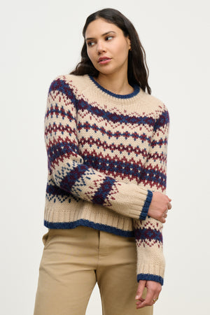 A woman stands against a plain background wearing the MAKENA ALPACA SWEATER by Velvet by Graham & Spencer, featuring a cozy cream Fair Isle design with blue and red patterns, paired with tan pants and her right arm crossed.