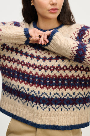A person in the MAKENA ALPACA SWEATER by Velvet by Graham & Spencer—beige and blue Fair Isle with maroon accents—raises one arm across their chest; their head is partially out of frame.