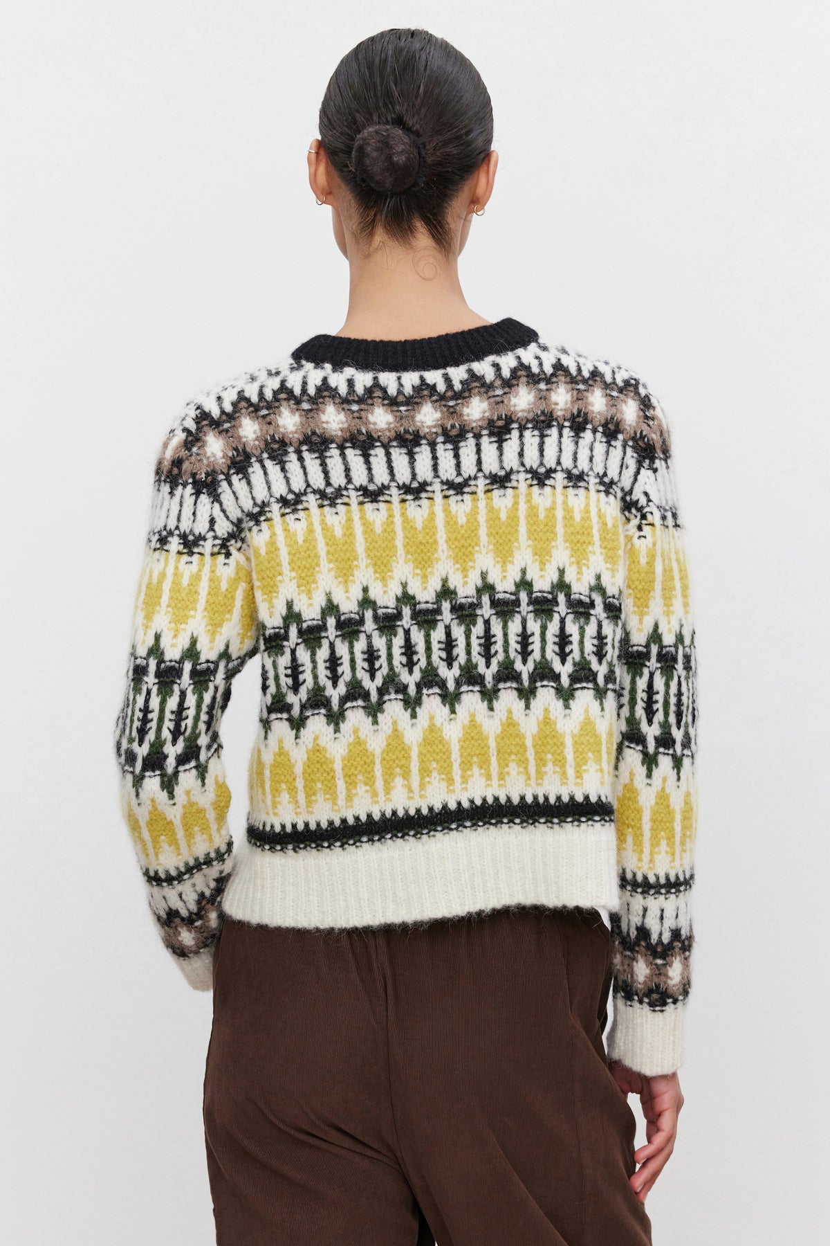 A person with a bun hairstyle is wearing the THELMA ALPACA FAIR ISLE SWEATER by Velvet by Graham & Spencer, featuring yellow, black, and white geometric designs, paired with brown pants, facing away from the camera against a plain background.-38373287329985