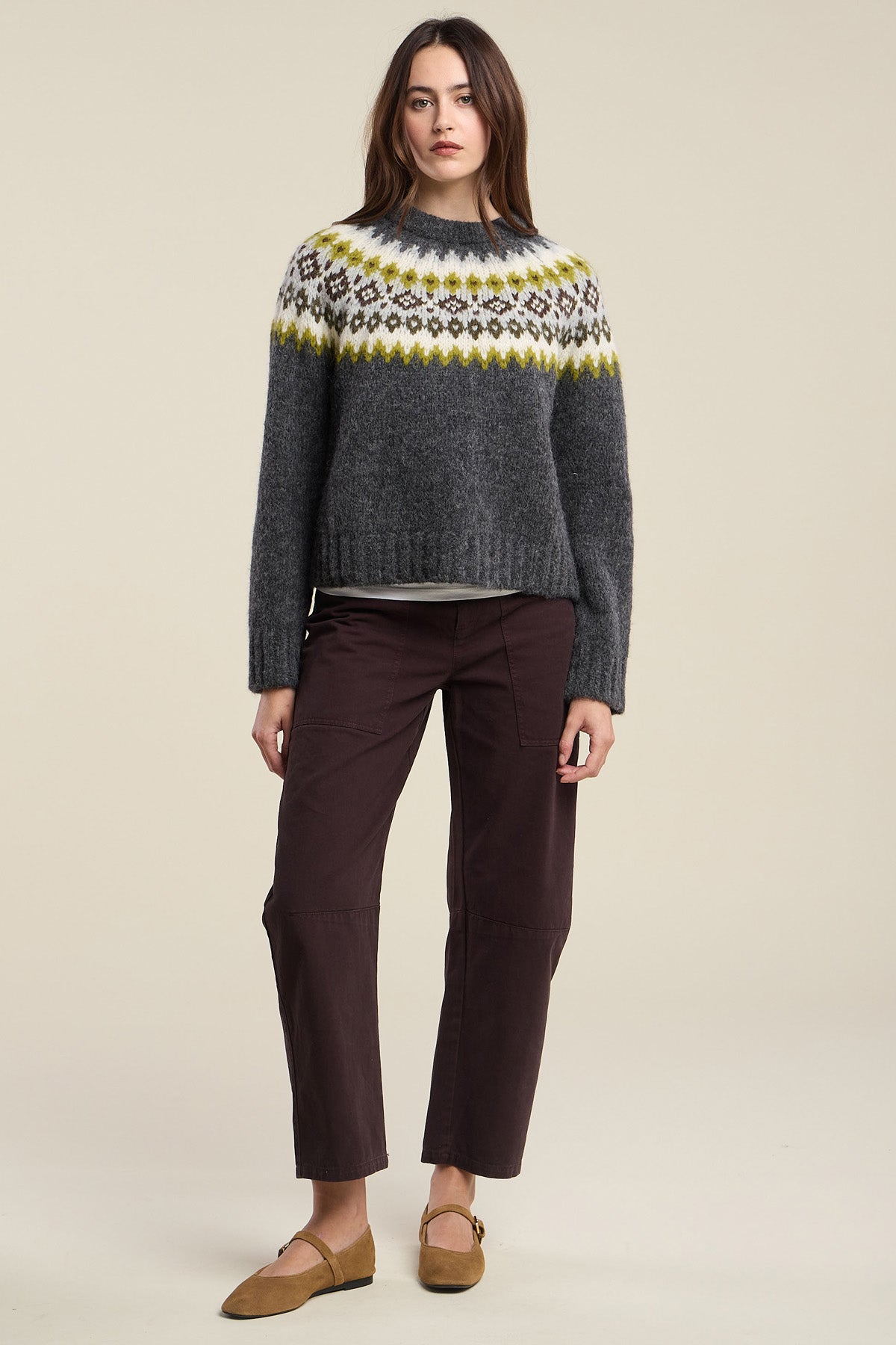 A woman stands against a plain beige background wearing the EDITH ALPACA SWEATER by Velvet by Graham & Spencer in a gray, yellow, and white Fair Isle pattern, paired with brown pants and tan flats.-42063737323713