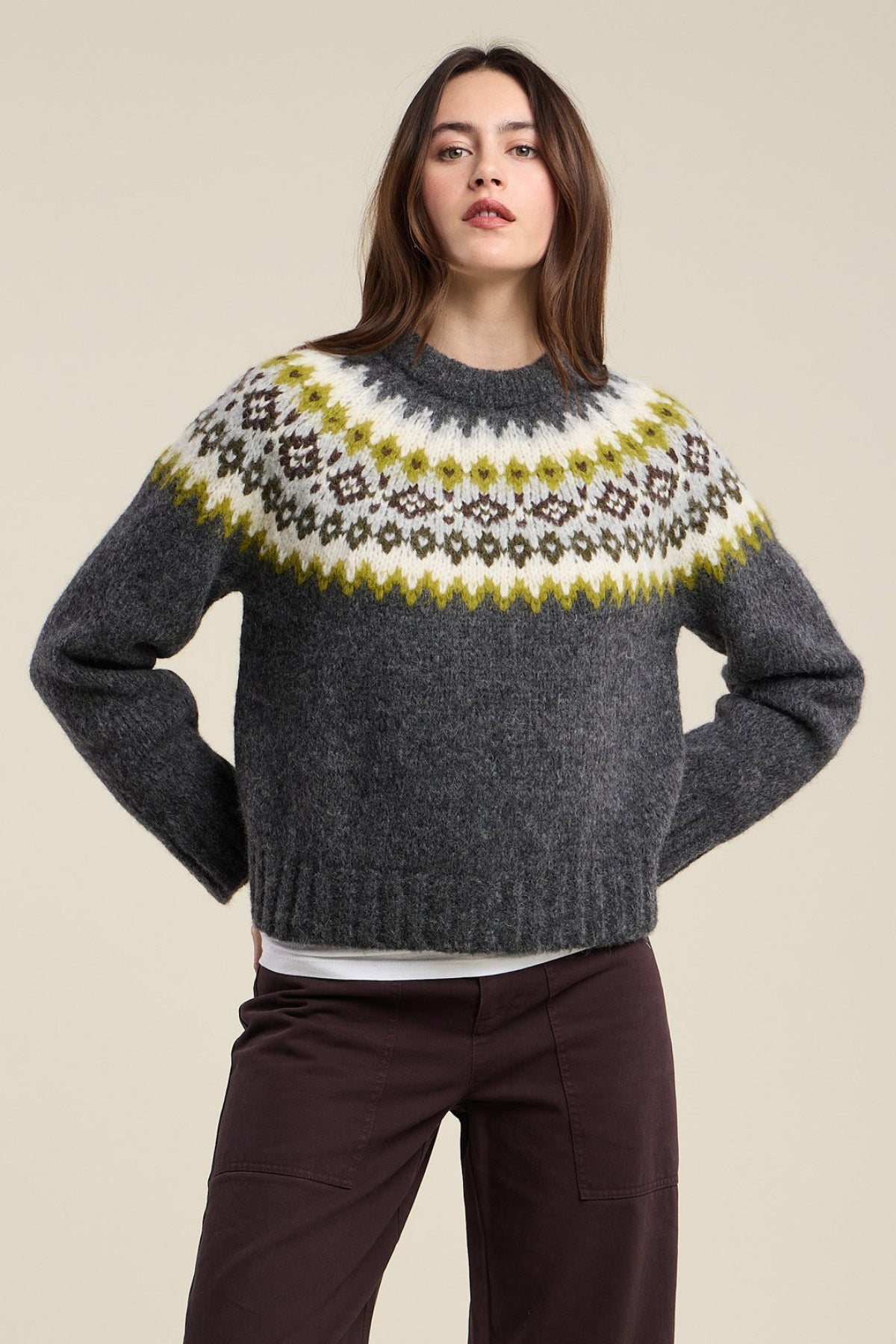 A woman wears the EDITH ALPACA SWEATER by Velvet by Graham & Spencer—a dark grey Fair Isle knit with a white, yellow, and brown patterned yoke—paired with dark brown pants, standing against a plain beige background.