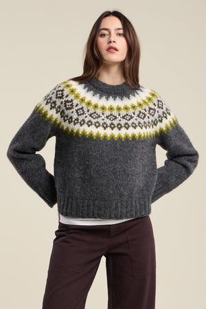 A woman wears the EDITH ALPACA SWEATER by Velvet by Graham & Spencer—a dark grey Fair Isle knit with a white, yellow, and brown patterned yoke—paired with dark brown pants, standing against a plain beige background.