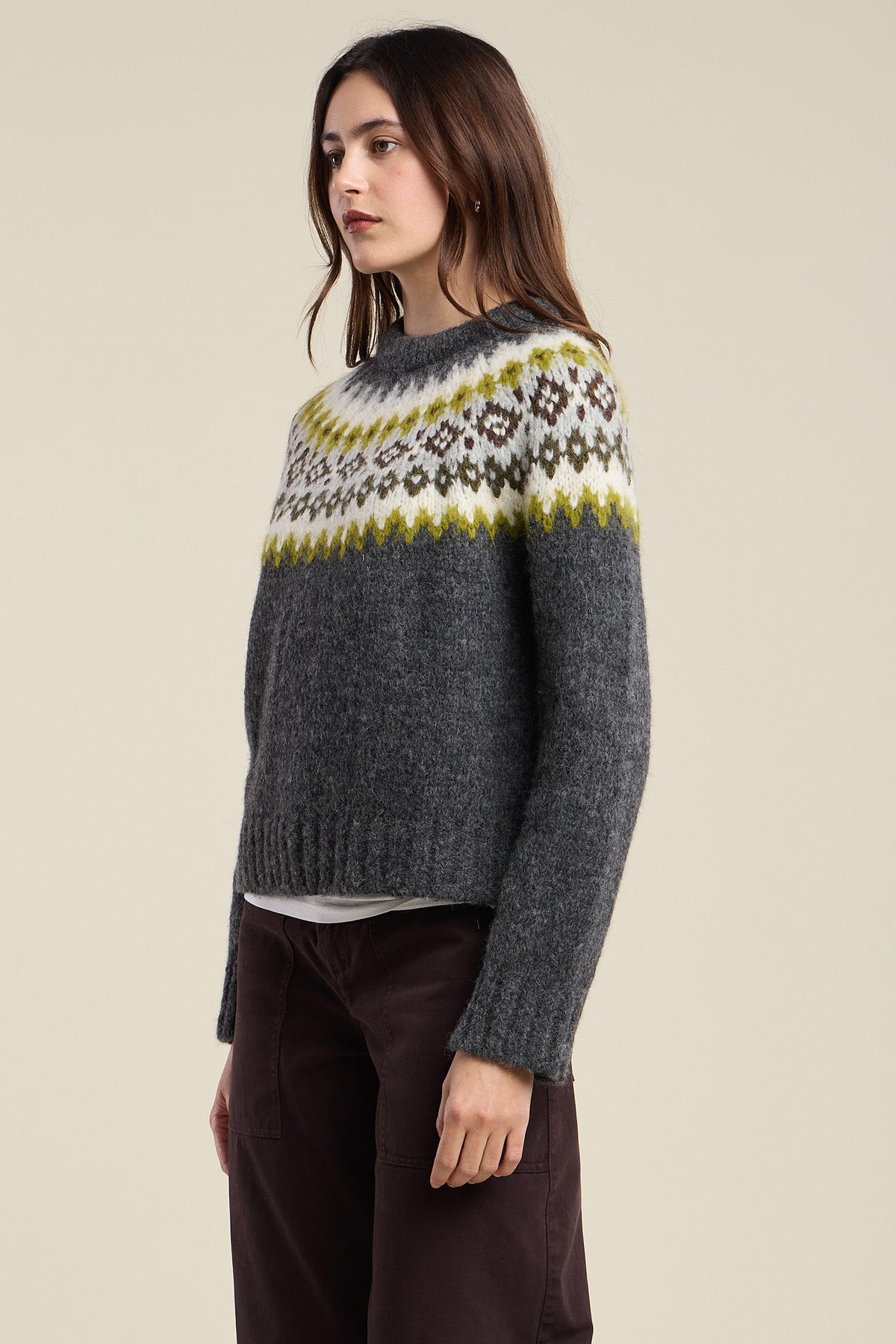 A woman stands against a plain background wearing the EDITH ALPACA SWEATER by Velvet by Graham & Spencer—a dark gray alpaca blend sweater featuring a white, brown, and yellow geometric pattern across the chest—paired with dark pants.