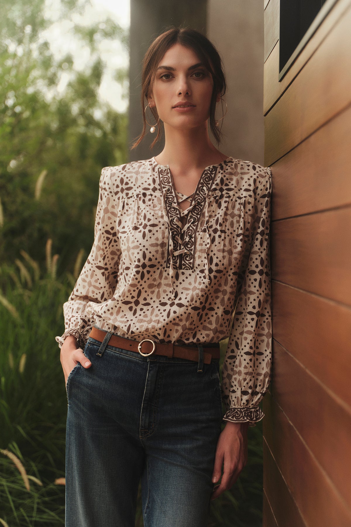   A woman wearing jeans and a blouse leaning against a wall, accessorized with COCO HOOP EARRINGS BY BYCHARI. 