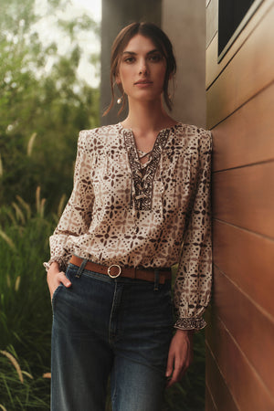 A woman wearing jeans and a blouse leaning against a wall, accessorized with COCO HOOP EARRINGS BY BYCHARI.