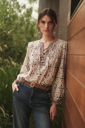 A woman wearing jeans and a AUDETTE PRINTED BOHO TOP by Velvet by Graham & Spencer leaning against a wall.