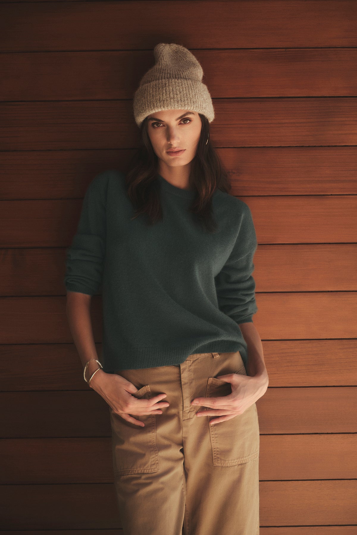 A woman in a green Velvet by Graham & Spencer Brynne Cashmere Crew Neck sweater and tan pants leaning against a wall.-26883604349121