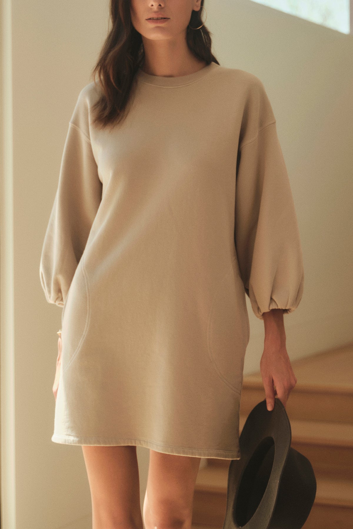   A person wearing a long-sleeve beige dress holds the CHELSEA RIBBON TRIM FEDORA by Velvet by Graham & Spencer, standing indoors near steps. 