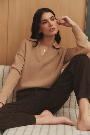 A woman is sitting on a couch wearing a LUCIE POLO SWEATER by Velvet by Graham & Spencer and black pants.