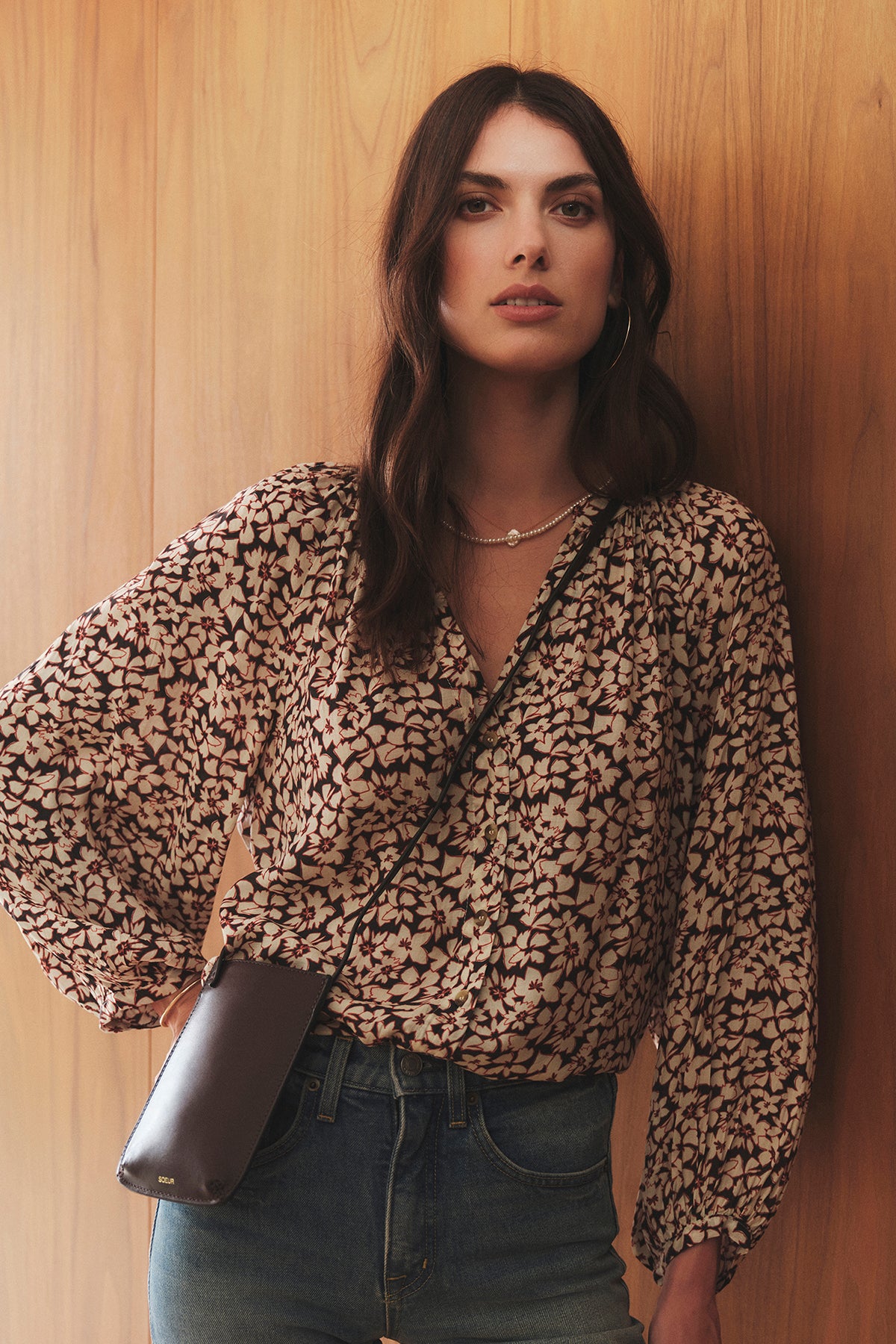   A stylish woman, representing the Soeur brand, wearing a blouse and jeans elegantly leans against a wall with the NOE Bag by Soeur in hand. 