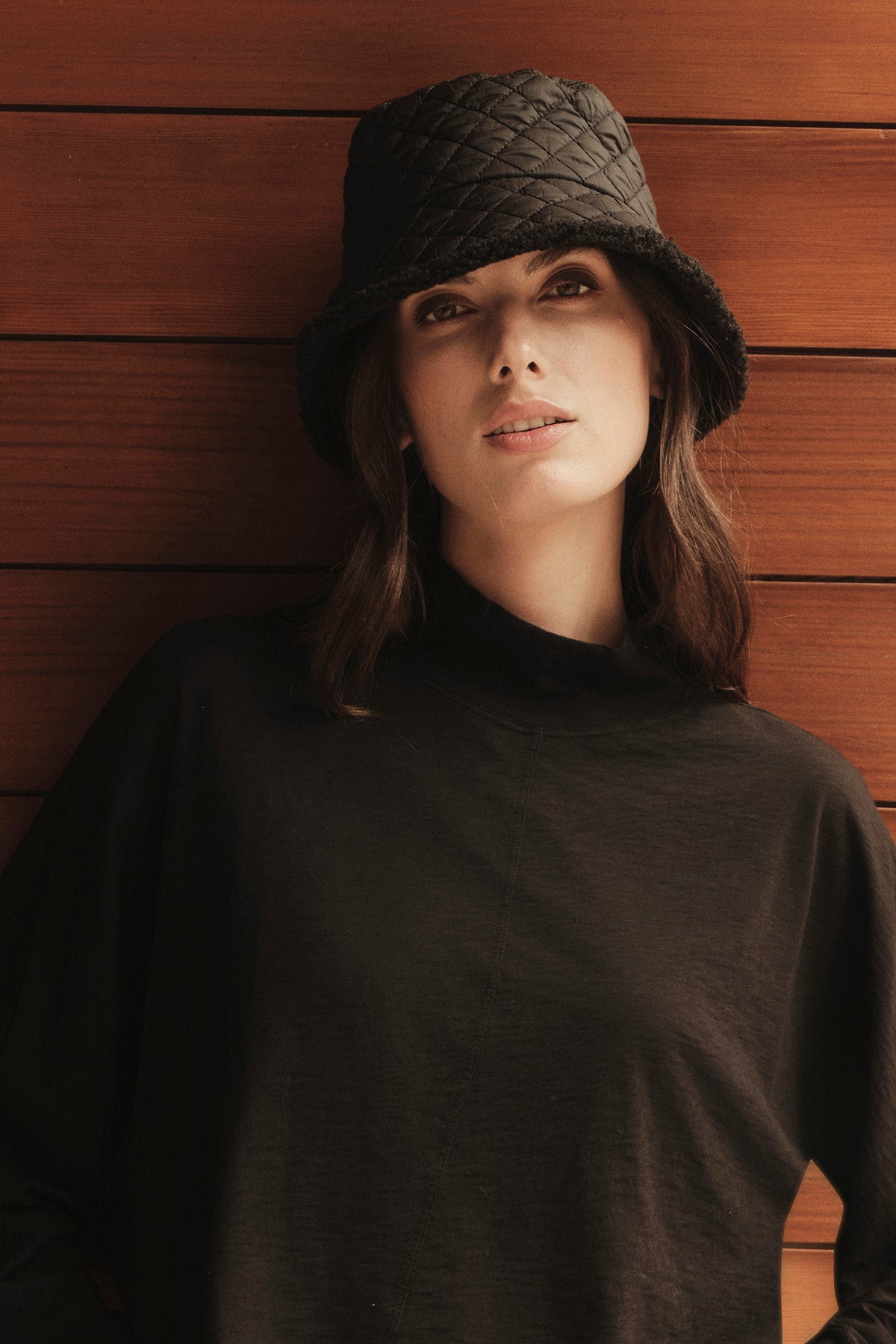   A woman wearing a black QUILTED BUCKET HAT by Velvet by Graham & Spencer with faux fur trim leaning against a wooden wall. 