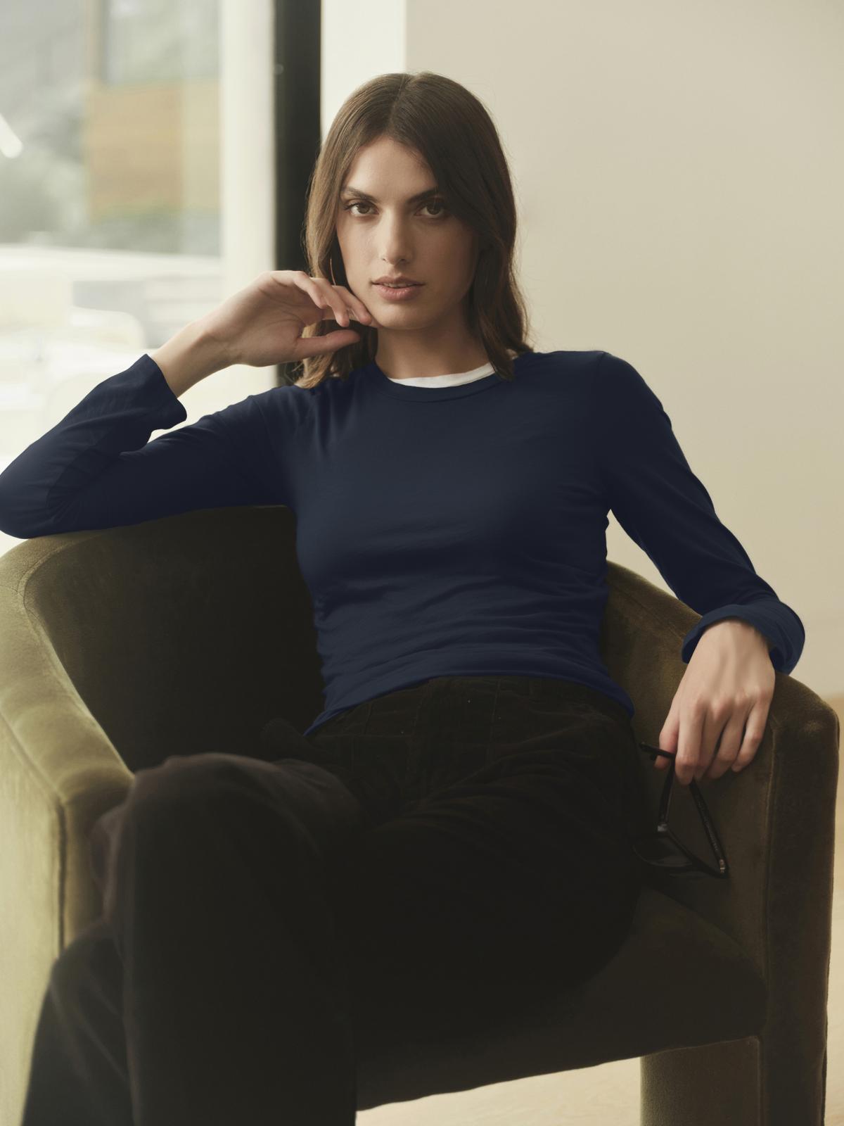   A woman wearing Velvet by Graham & Spencer's ZOFINA GAUZY WHISPER FITTED CREW NECK TEE, a navy long-sleeved shirt, sitting in a chair. 