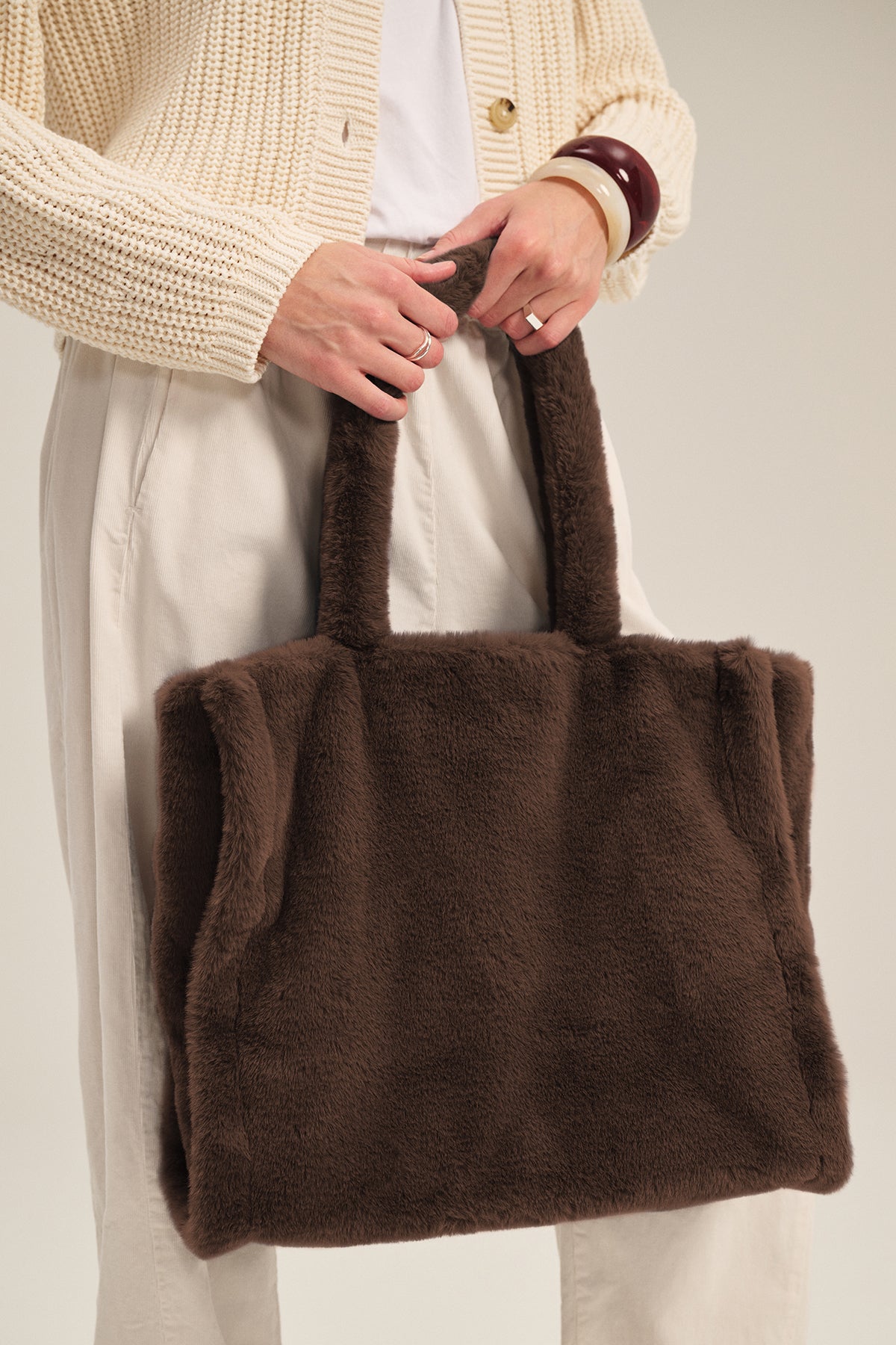 A person in a cream sweater and light pants holds the LIBBY LUXE FAUX FUR TOTE by Velvet by Graham & Spencer—a spacious, cozy accessory ideal for winter outings.