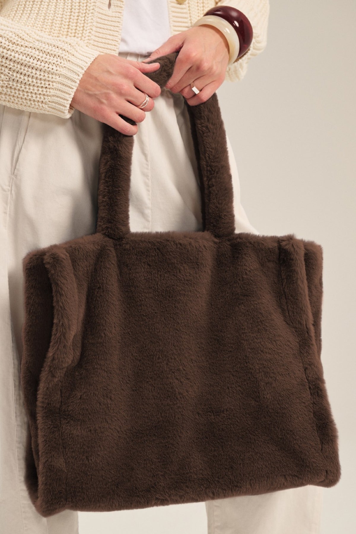 A person holds the LIBBY LUXE FAUX FUR TOTE by Velvet by Graham & Spencer, featuring two handles—an ideal cold-weather accessory—paired with light-colored pants and a textured sweater.-40341041283265