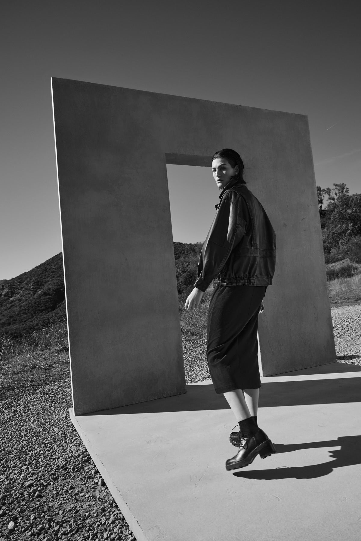   A person wearing the CORONADO DRESS by Velvet by Jenny Graham stands on a concrete platform next to a rectangular structure in an outdoor setting. The image is in black and white. 