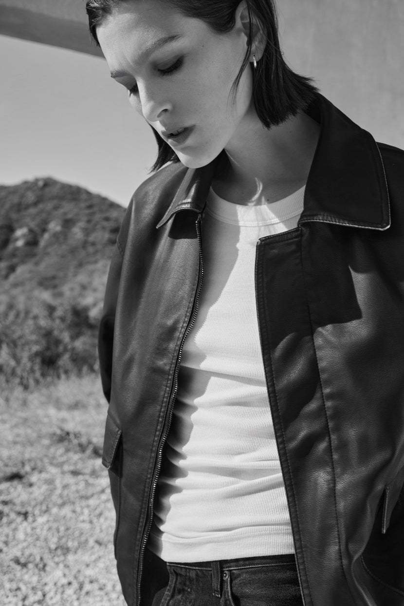 A person wearing a Velvet by Jenny Graham CALIFORNIA FAUX LEATHER JACKET over a white top stands outdoors with their head tilted slightly downward, set against a hilly landscape in an evocative black and white image.