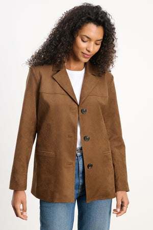 A woman wears the SALIE FAUX SUEDE JACKET by Velvet by Graham & Spencer over a white shirt and blue jeans, standing against a plain background while looking down and smiling slightly.