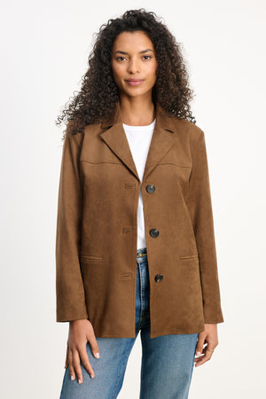 A woman wears the SALIE FAUX SUEDE JACKET by Velvet by Graham & Spencer in brown over a white shirt and blue jeans, standing against a plain light background.