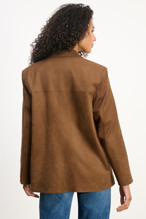 A person with curly hair wears the SALIE FAUX SUEDE JACKET by Velvet by Graham & Spencer, featuring a notched collar in brown, paired with blue jeans, facing away from the camera against a plain white background.