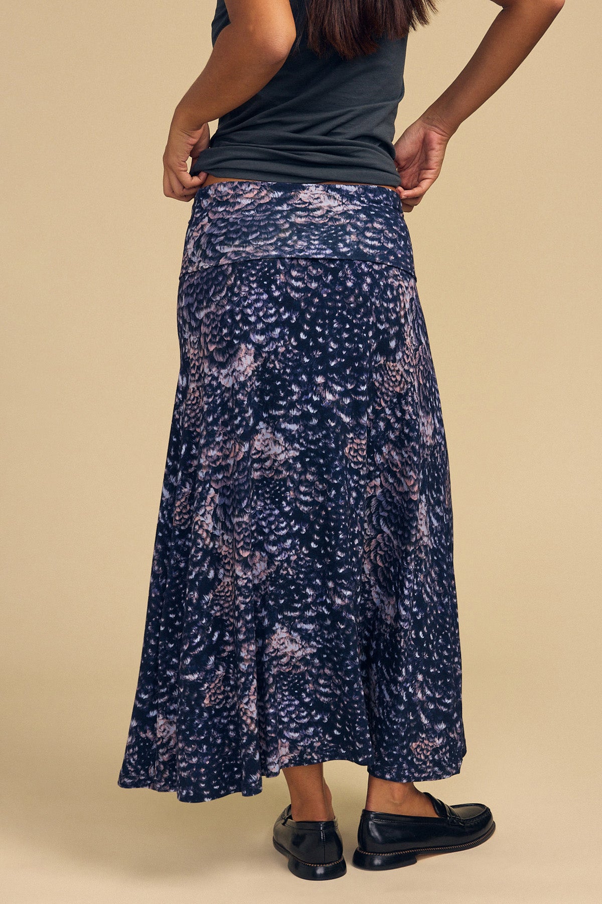 A woman viewed from behind wears a dark Velvet by Graham & Spencer VALERIE PRINTED SKIRT, a knee-length column maxi skirt with an abstract feather-like print, a dark t-shirt, and black loafers.-46489759645889