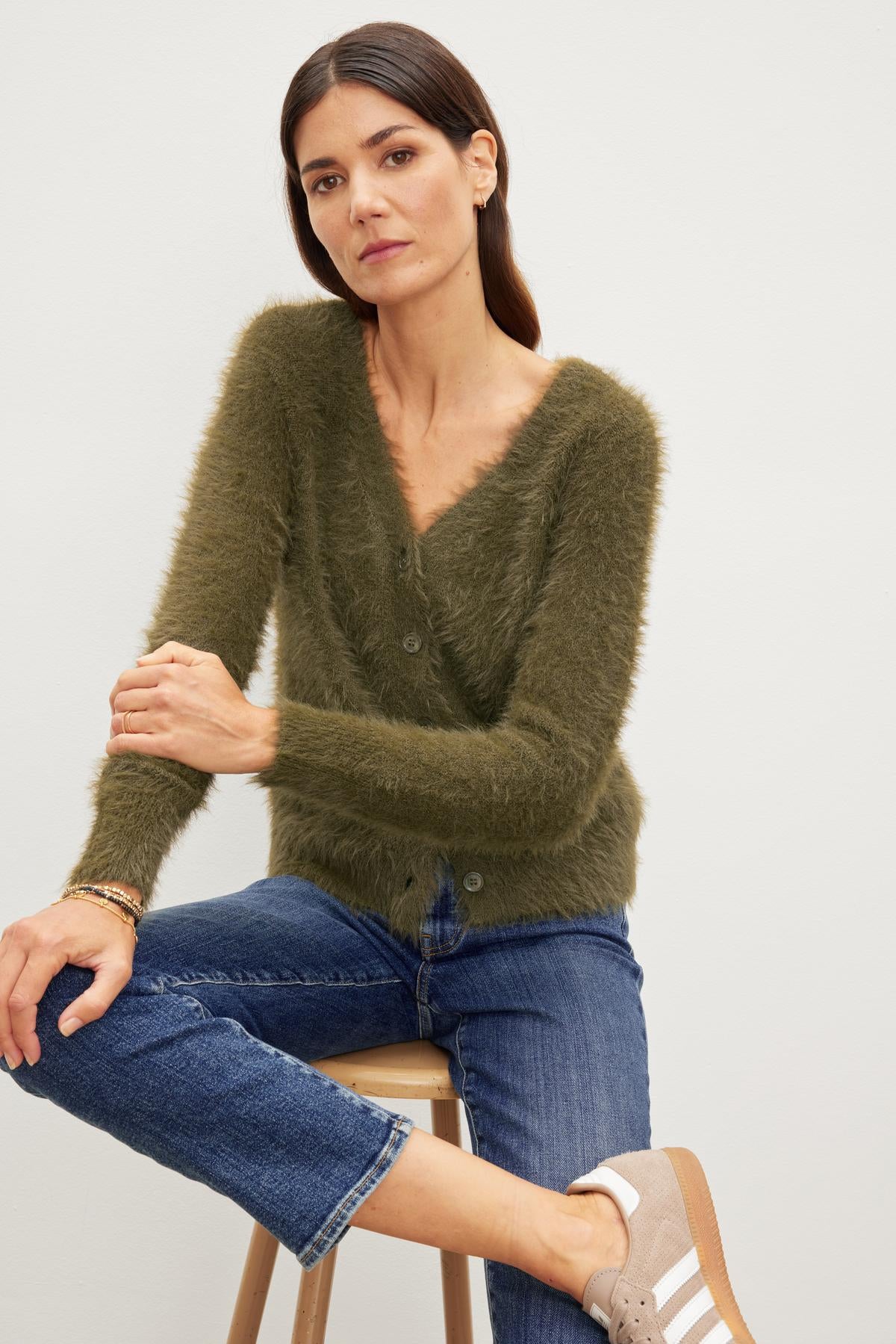 A woman is sitting on a stool wearing jeans and a soft green ELLE FEATHER YARN CARDIGAN made by Velvet by Graham & Spencer.-35630370488513