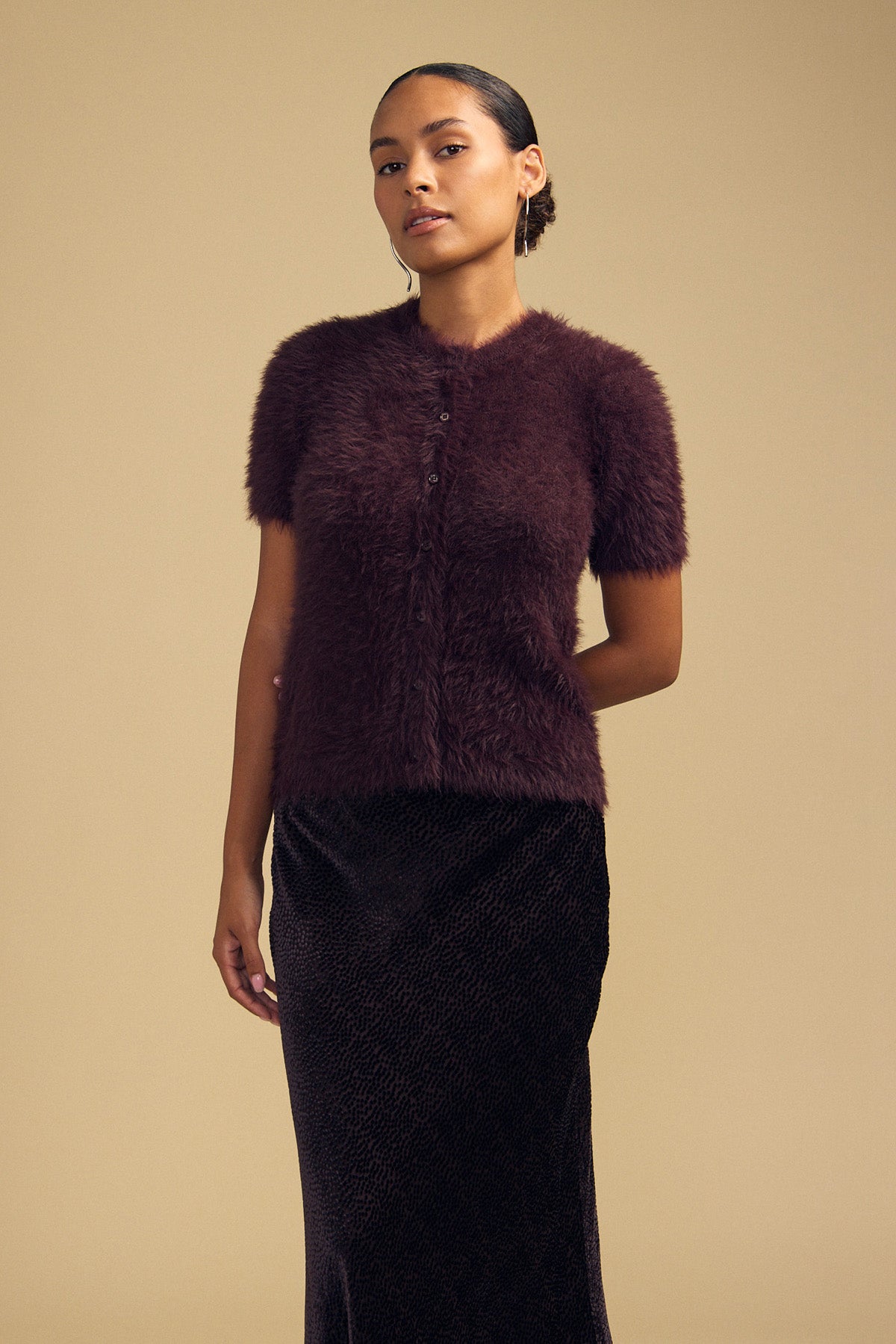 A person wears a short-sleeved, fuzzy dark purple GABBY FEATHER YARN CARDIGAN in lightweight feather yarn and a dark patterned midi skirt.